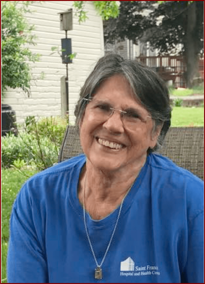 A smiling woman with glasses, wearing a blue t-shirt with a health center logo, sitting outdoors.