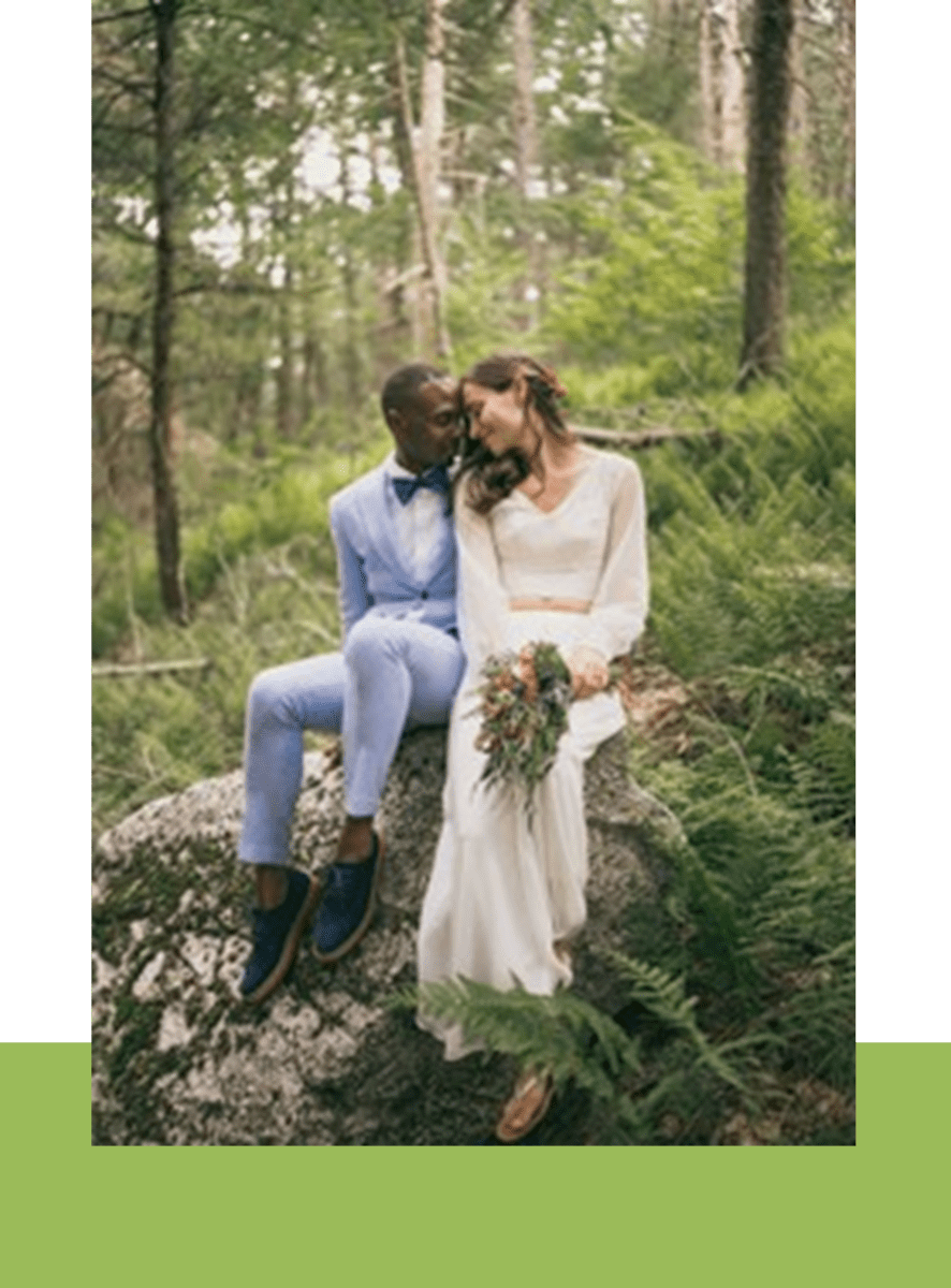 A bride and groom smiling at each other while sitting on a large rock in a lush forest. the bride holds a bouquet, and the groom wears a light blue suit.