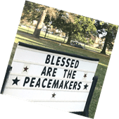 A sign with the message "blessed are the peacemakers" surrounded by stars, displayed in a park setting.