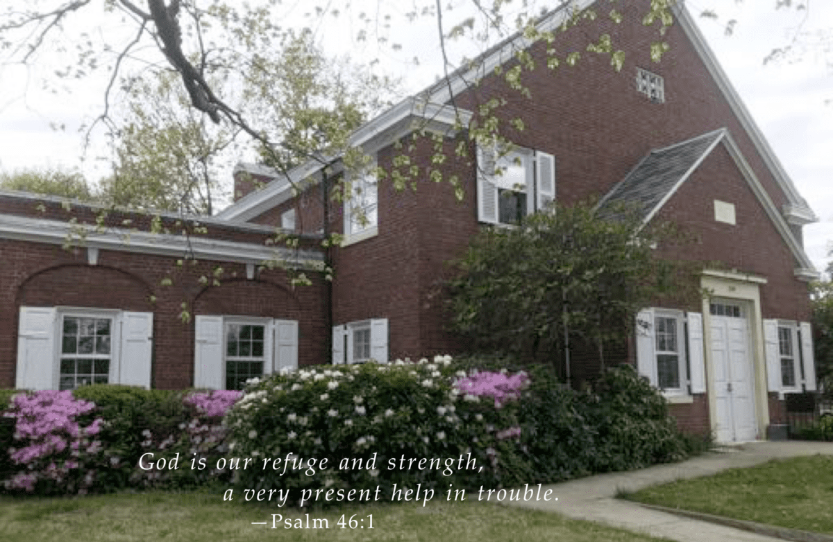 A quaint brick building with blooming bushes and a biblical verse, psalm 46:1, displayed on the front lawn.