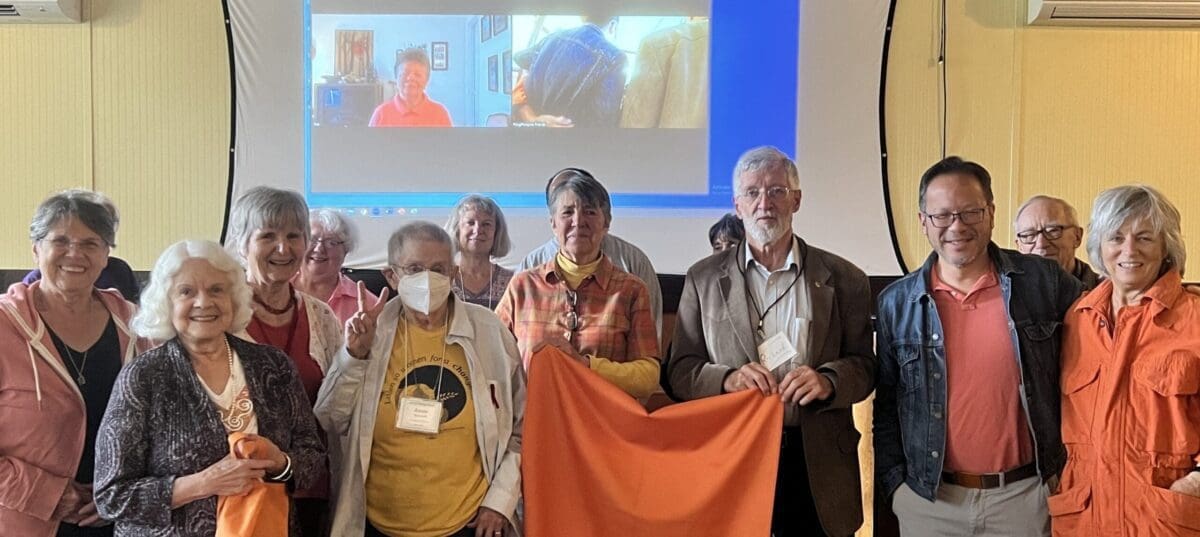 A group of people, some holding an orange cloth, pose for a photo in front of a projection screen displaying a video call. One person wears a face mask.