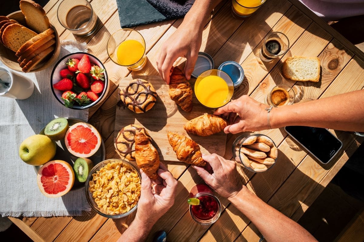 Four people reaching for croissants on a table set with breakfast items including juice, fruit, cereal, bread, and pastries.