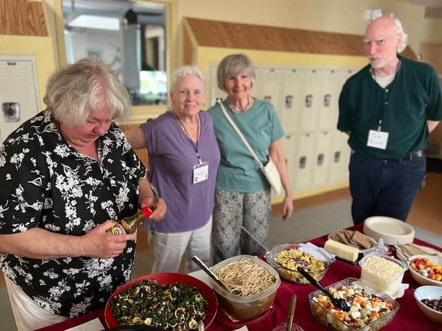 Four elderly individuals stand at a table full of various dishes, with one person serving themselves from a salad bowl in a casual setting.