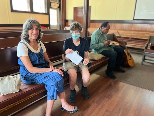 Three people are seated on wooden benches. The first two, both women, are conversing. Behind them, a man is eating with a guitar next to him. One woman is holding an envelope, the other is wearing a face mask.