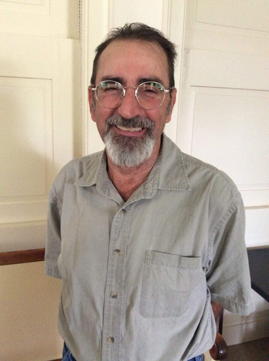 Rosario, a man with glasses and a grey beard is smiling, wearing a light grey collared shirt, standing in a room with white paneled walls.