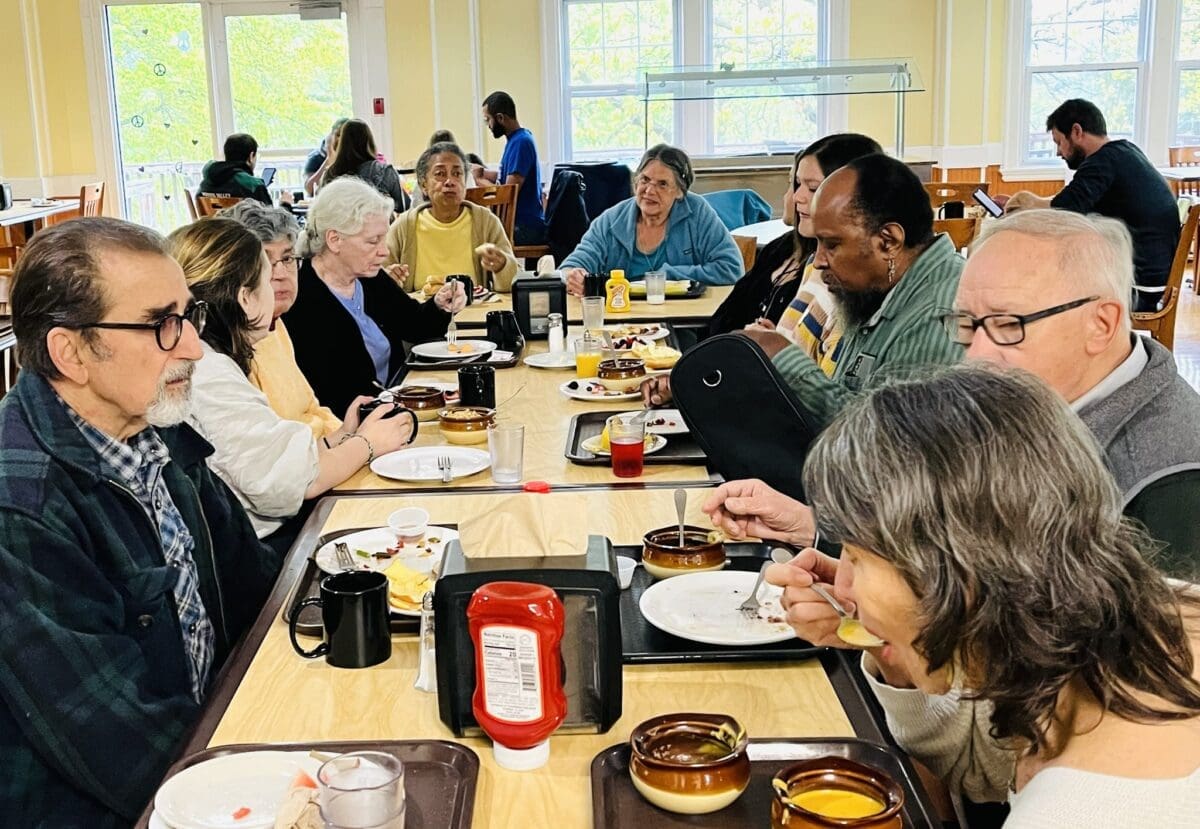 A group of people, seated around a table in a dining area, are eating and chatting. Plates, bowls, and condiments are visible on the table.