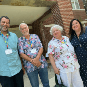 Four people stand outside a brick building, wearing name tags and colorful lanyards, posing for a photo and smiling.