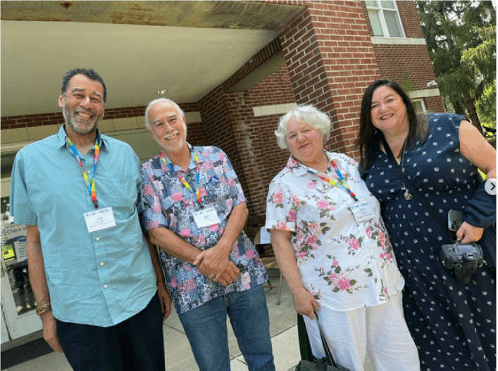 Four people stand outside a brick building, wearing name tags and colorful lanyards, posing for a photo and smiling.