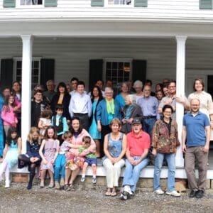 A group of about 30 people, including adults and children, pose on the porch and steps of a white house with green shutters. Some are standing while others are seated.