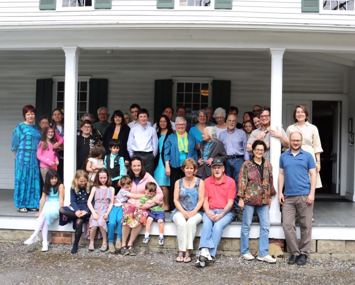 A group of about 30 people, including adults and children, pose on the porch and steps of a white house with green shutters. Some are standing while others are seated.