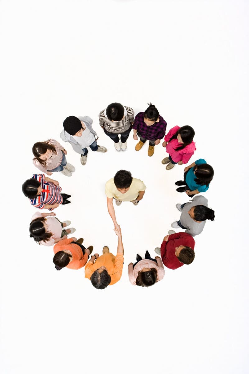 A group of people stands in a circle facing inward, with two individuals in the center shaking hands. The photo is taken from an overhead perspective.