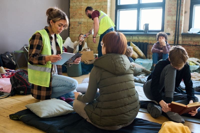 A group of people in a room with exposed brick walls, some seated on the floor reading and talking, while others stand, all wearing casual clothes and some in reflective safety vests.