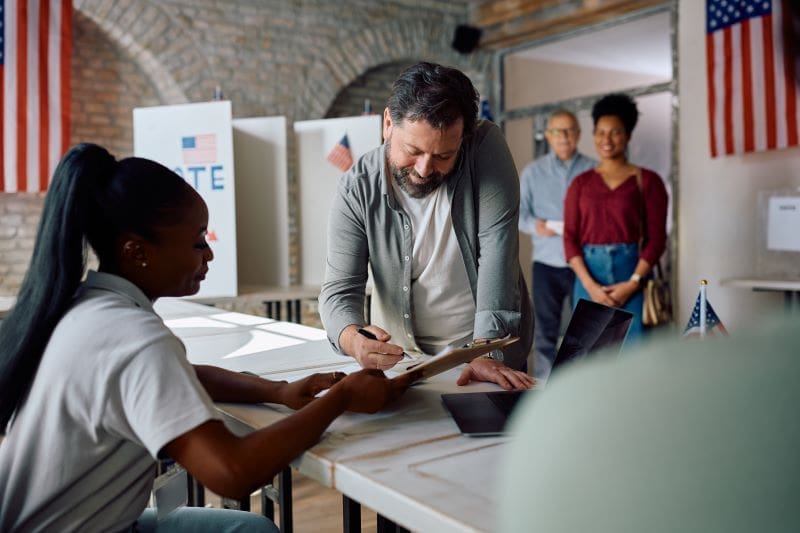 A man signs a document at a voting station while an official looks on. Two American flags are visible in the background, and two people wait in line behind him.