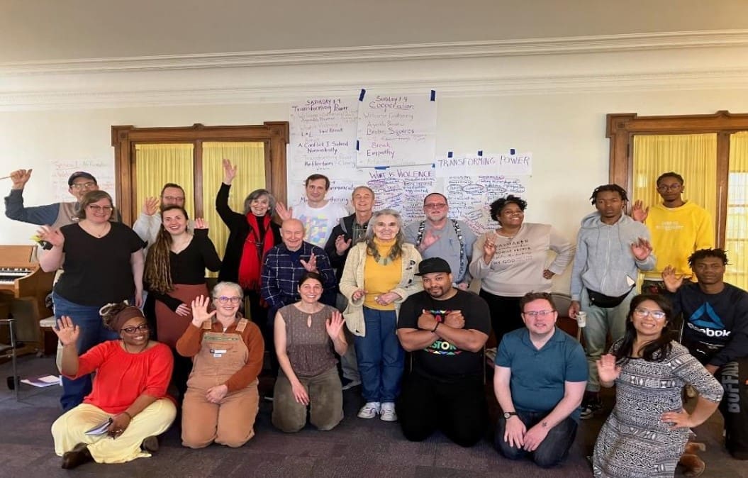 A group of 19 people standing and sitting in a room, some raising their hands. They are in front of whiteboards with notes. The atmosphere appears casual and friendly.