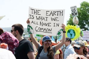 People are participating in a rally. A woman in the center holds a sign that reads, "The earth is what we all have in common," with images of the globe and other signs visible around her.