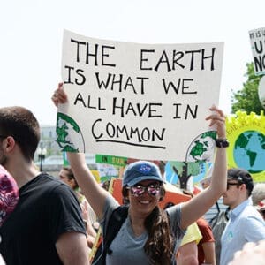 People are participating in a rally. A woman in the center holds a sign that reads, "The earth is what we all have in common," with images of the globe and other signs visible around her.