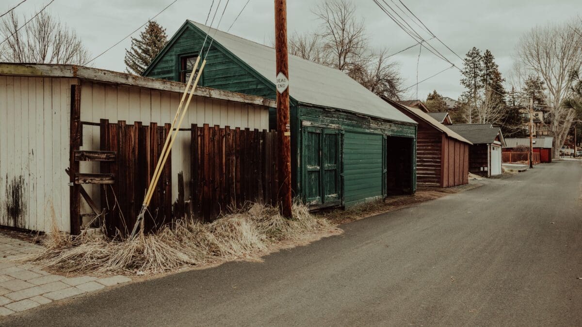 A narrow paved road lined with rustic sheds and buildings, some with peeling paint. Power lines stretch overhead, and the area has a quiet, almost deserted feel. Trees in the background.