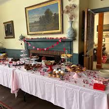 A long table covered with a white cloth is set with various pastries and cupcakes. Pink heart decorations are strung above a fireplace, and artwork hangs on the wall behind the display.